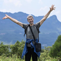 Man with portable oxygen concentrator connecetd and arms outstretched in a natural setting with mountains and greenery