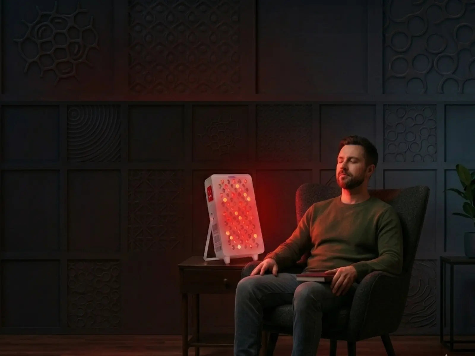 Man sitting in a chair with a red light therapy lamp in a dimly lit room.