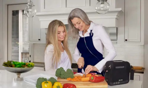 Two women in a kitchen preparing food with a portable oxygen concentrator on the counter.